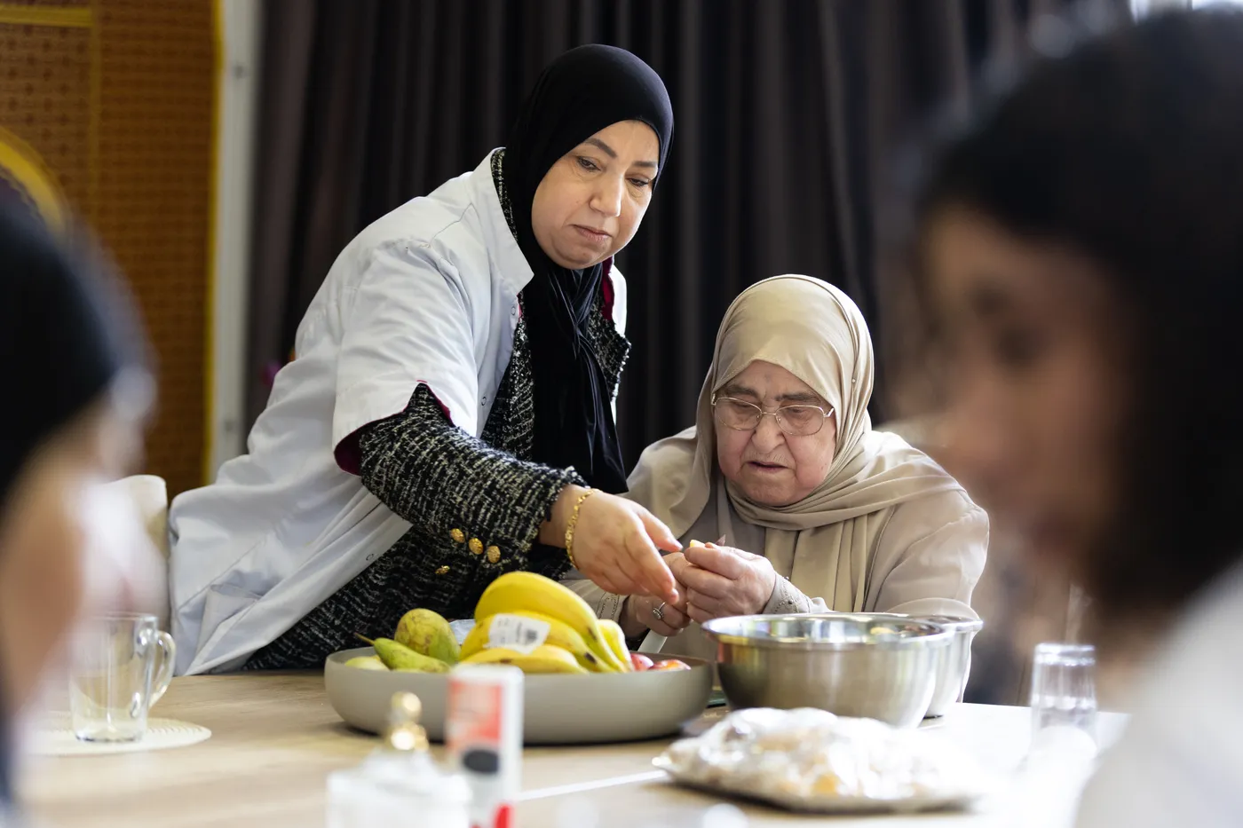 Een zorgverlener helpt een oudere vrouw tijdens een gezamenlijke activiteit aan tafel